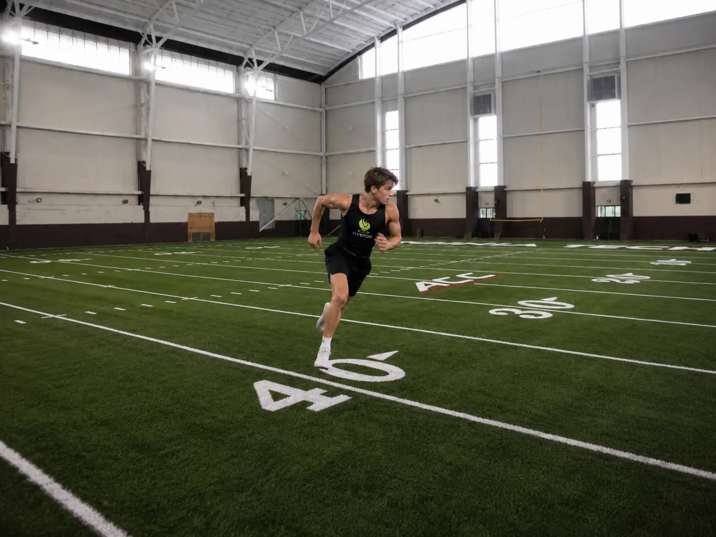 An athlete in a FiyrPod training top sprinting across the turf of an indoor training facility.